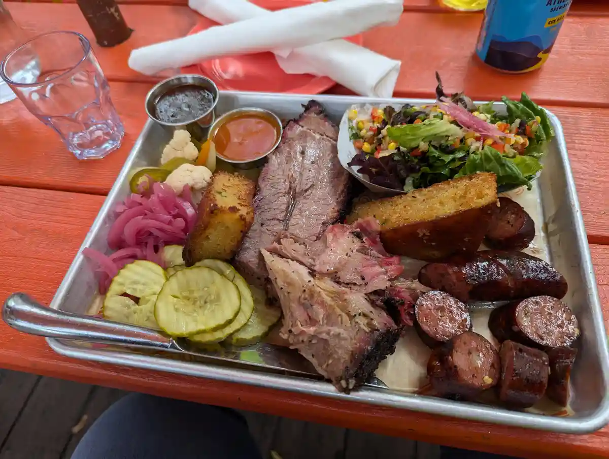 A metal tray holds a large, messy barbecue platter with sliced brisket, sliced sausages, cornbread, pickles, pickled onions…