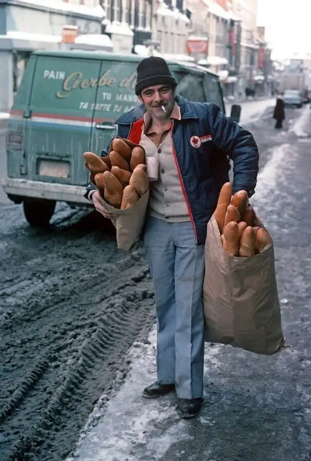A man in a winter hat and jacket stands on a snowy street holding two large sacks overflowing with baguettes. He has a…