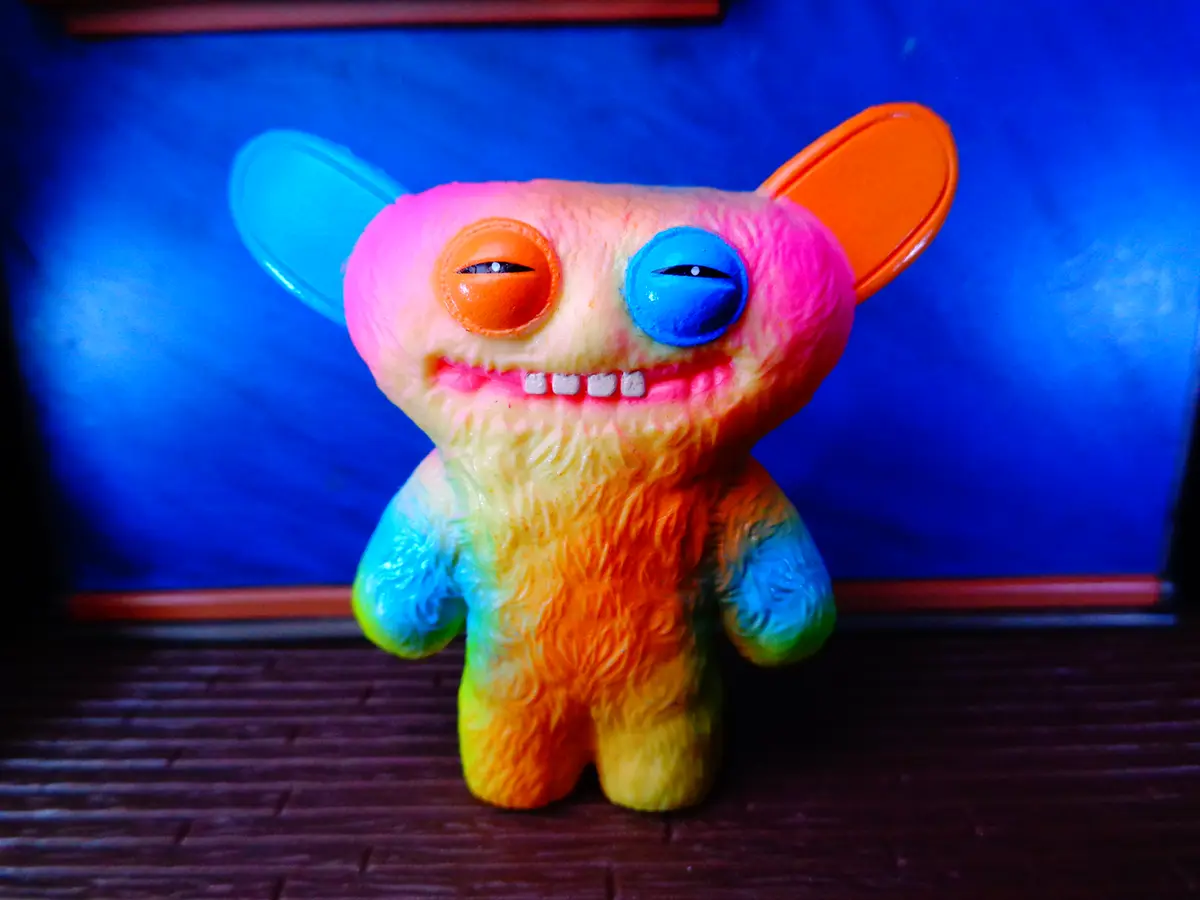 A colorful, fuzzy toy monster with mismatched orange and blue eyes, one ear blue and one orange, standing against a blue…