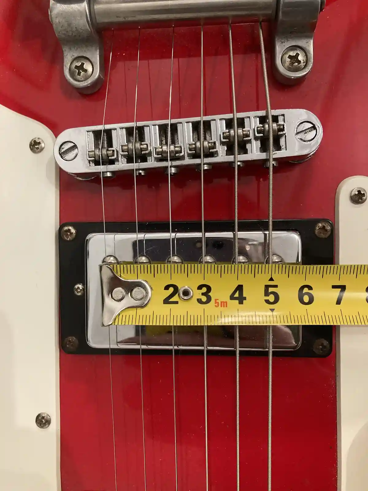 A close-up photo of a red electric guitar's bridge and pickup, with a yellow measuring tape stretched horizontally across…