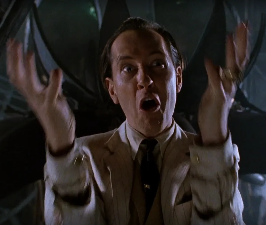 A man in a light-colored suit and dark tie, with a shocked and frantic expression, raises both hands in surrender or denial…