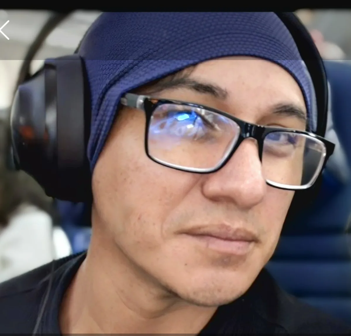 A close-up of a man wearing a blue head covering, black-rimmed glasses, and large black headphones, looking slightly…