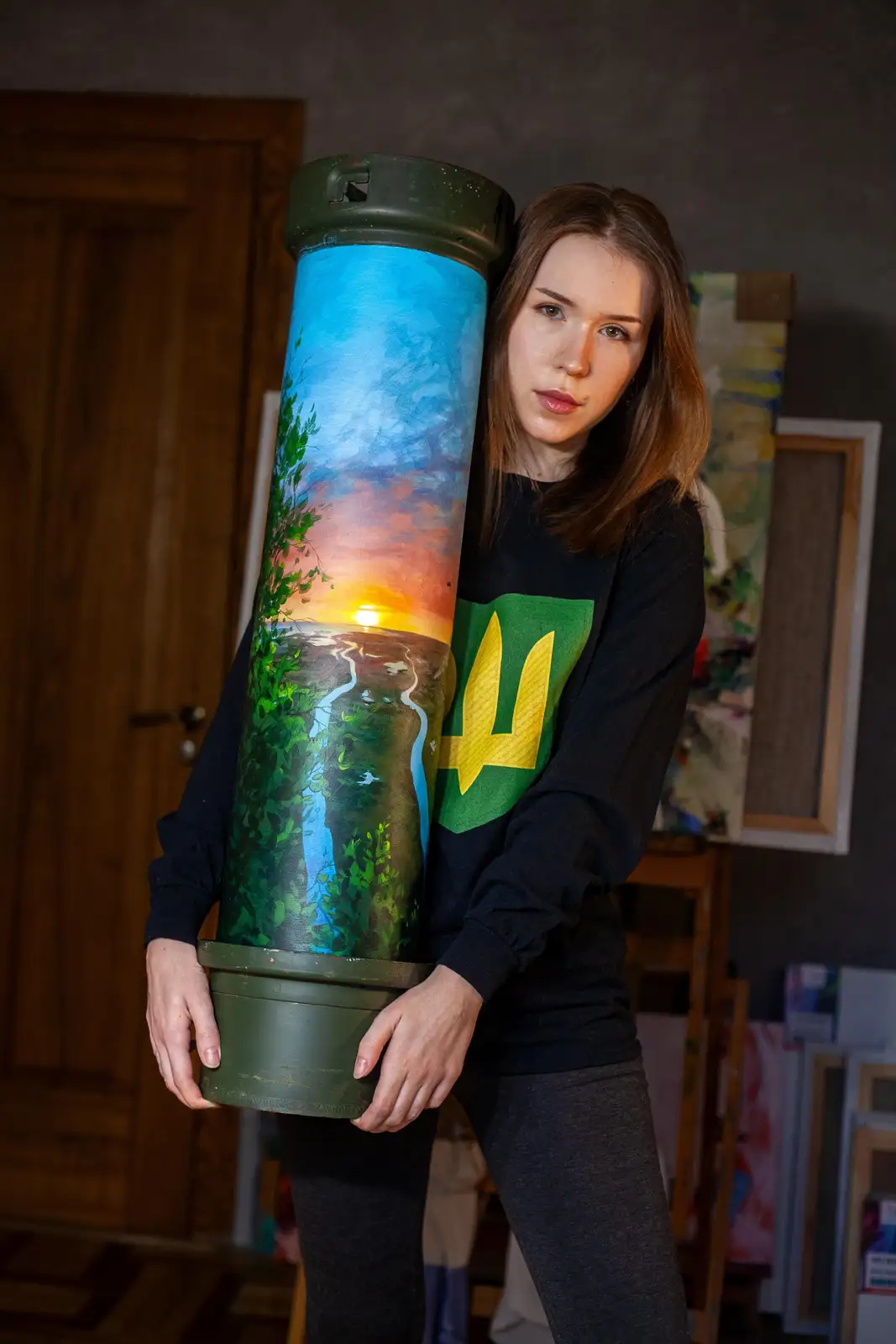 A woman in a black sweatshirt with a yellow and green Ukrainian trident (tryzub) emblem stands indoors holding a large…