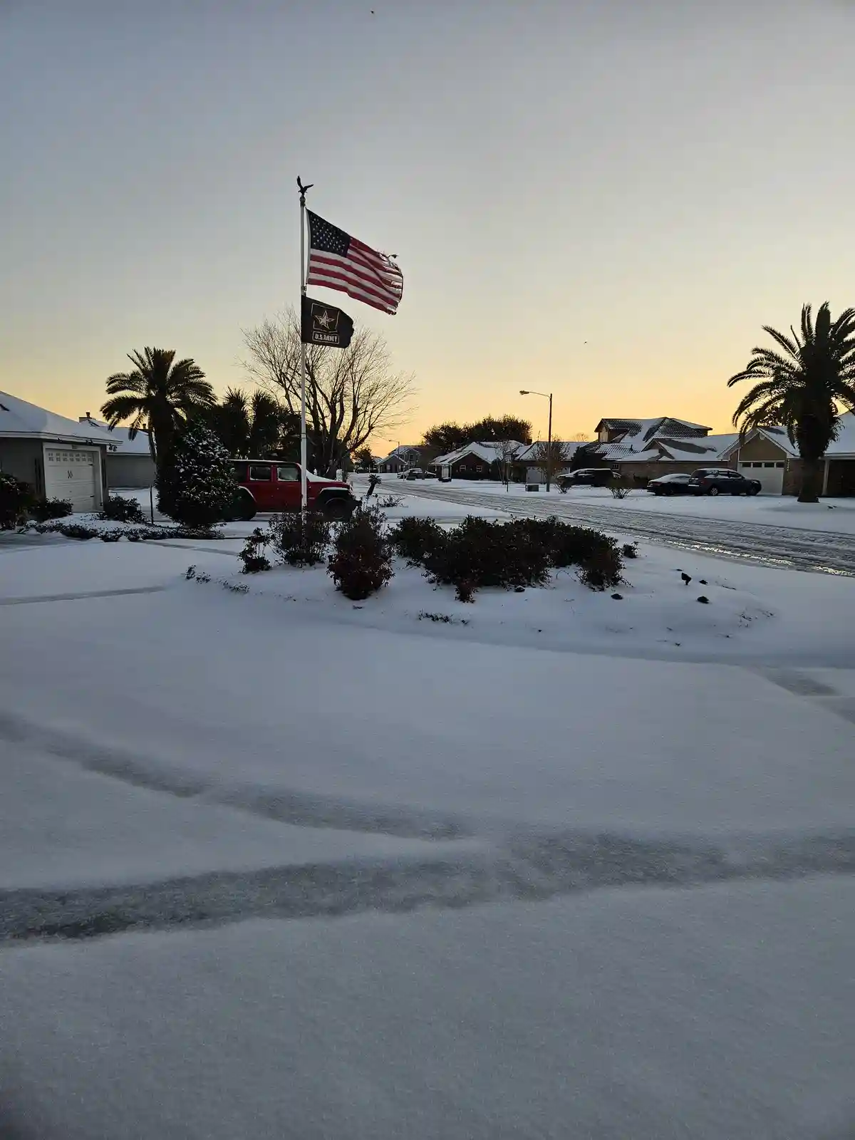 A snowy suburban street in Texas at sunset, with palm trees covered in snow, houses with snow on roofs, and an American flag…