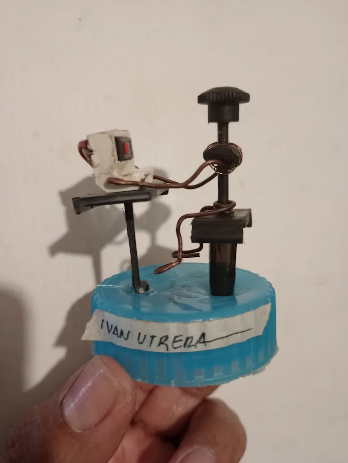 A hand holds a small, homemade-looking device built on a blue plastic bottle cap base. The contraption includes a red button…