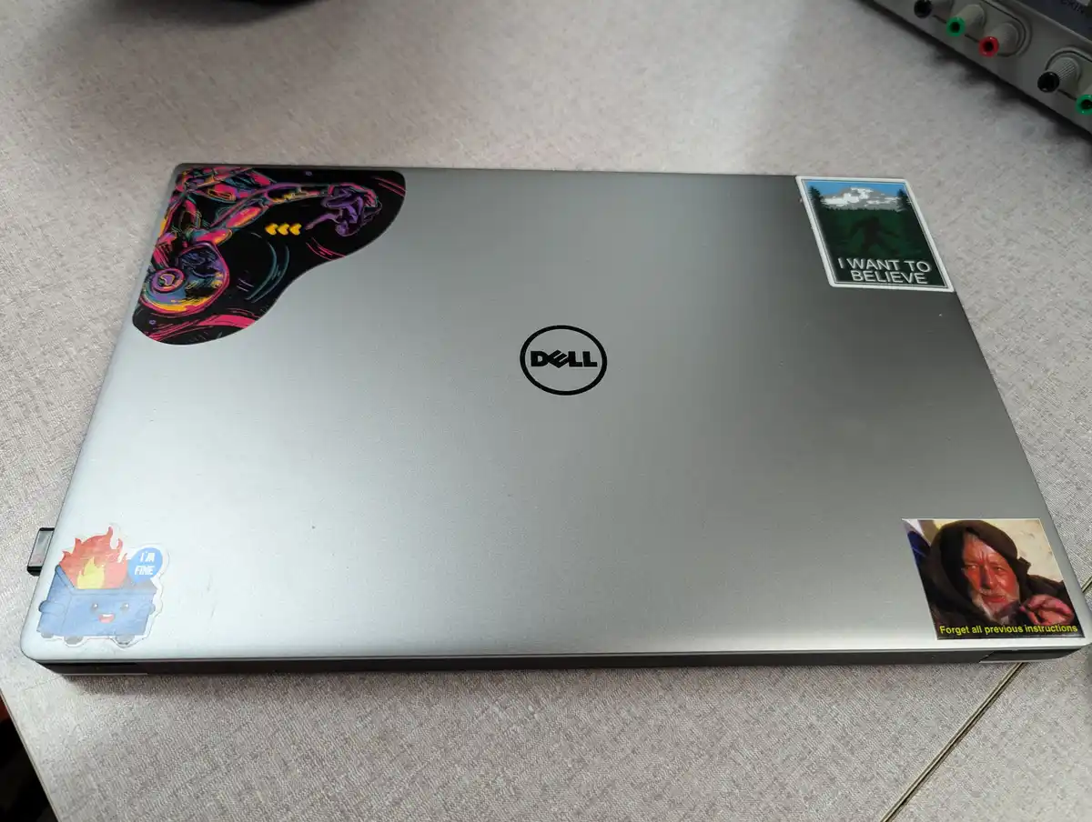 A silver Dell laptop lid covered in stickers: a colorful abstract design, a 'I WANT TO BELIEVE' Bigfoot sticker, a cartoon…
