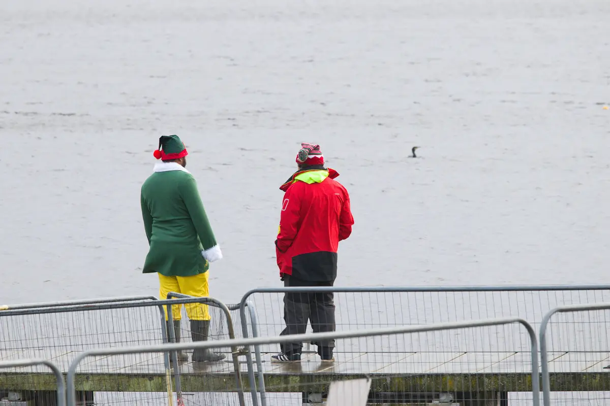 Two men stand on a wet pier overlooking a vast, flat, grey expanse of water. One wears a full green elf costume with yellow…