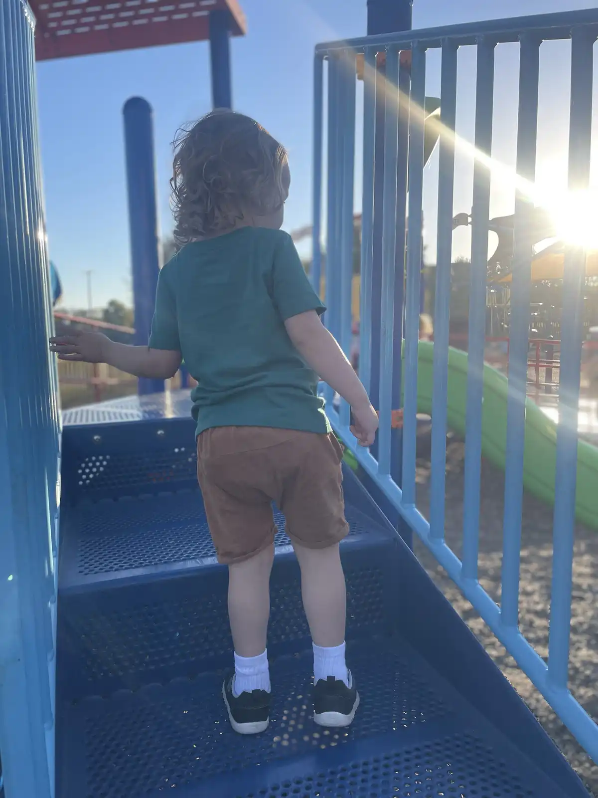 A young child with curly hair, wearing a green t-shirt, brown shorts, white socks, and black shoes, stands on a blue metal…
