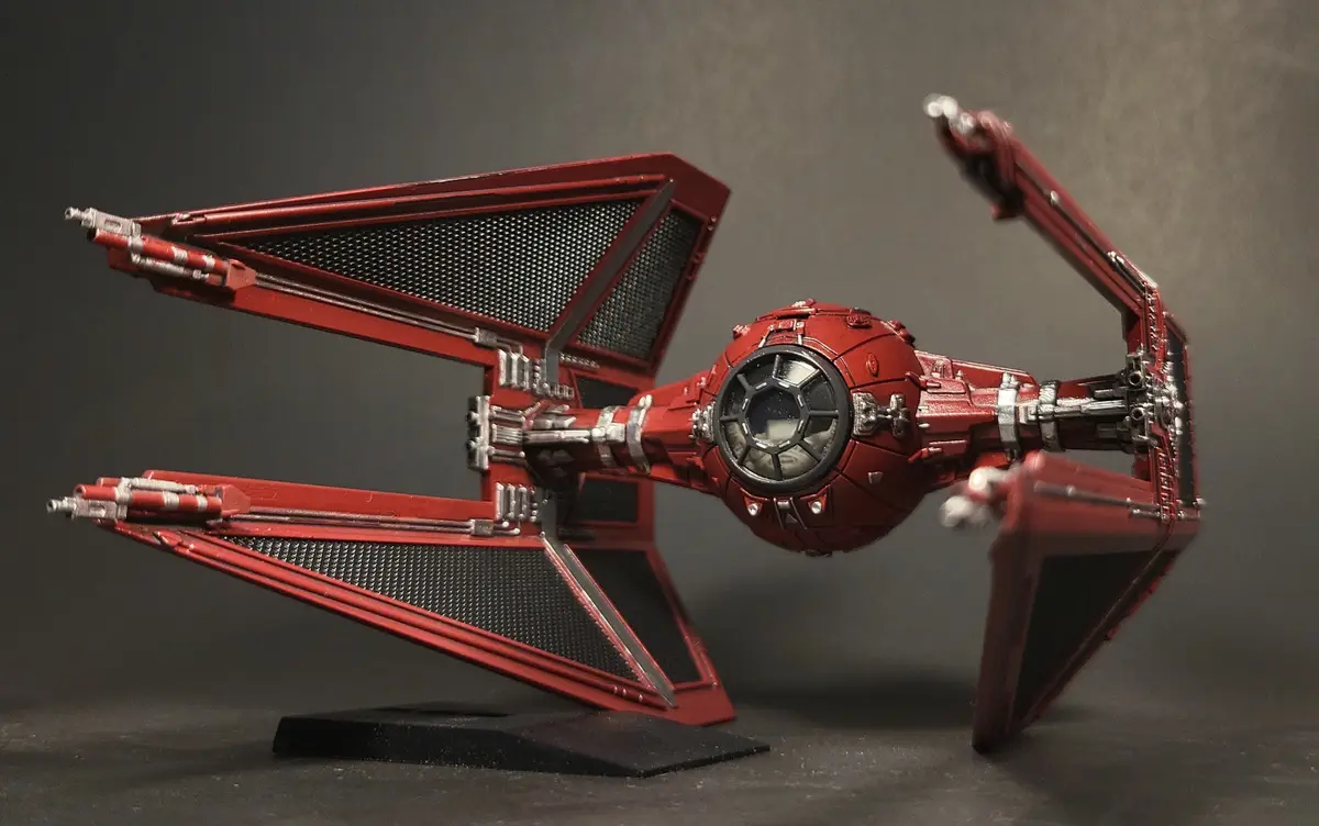 A detailed red model of a TIE fighter from Star Wars, displayed on a black stand against a dark gray background. The text…