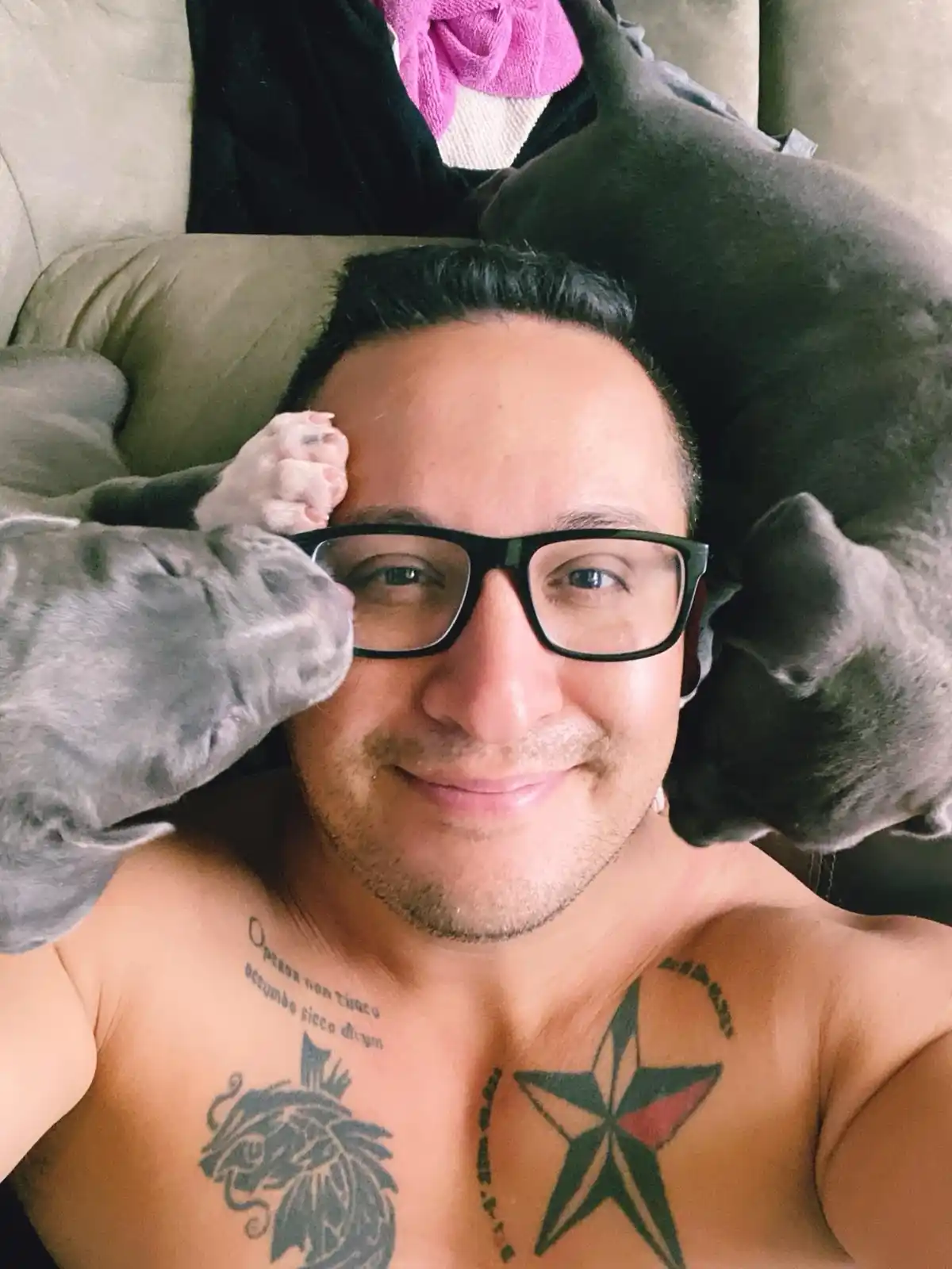 A shirtless man with glasses and chest tattoos lies on a couch, smiling as two grey dogs rest their heads on his shoulders…