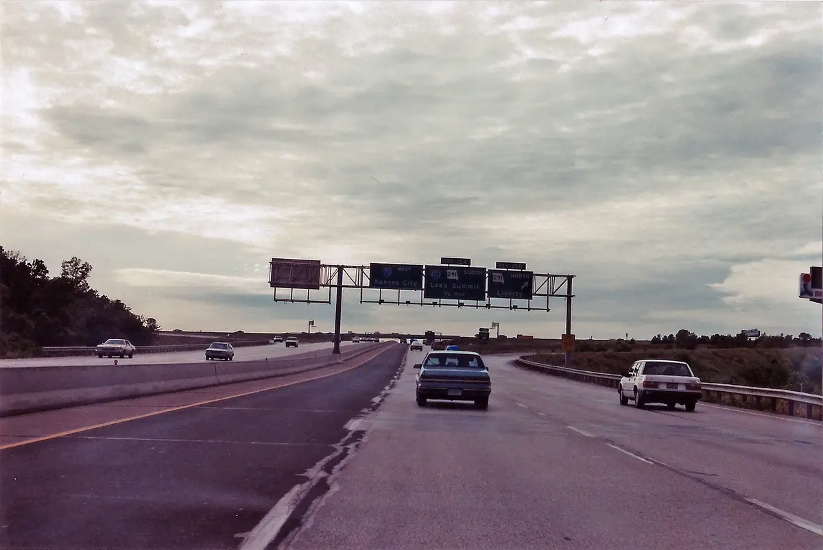 A vintage photo of a highway with multiple lanes and overhead road signs pointing to Kansas City, Lee's Summit, and Liberty…