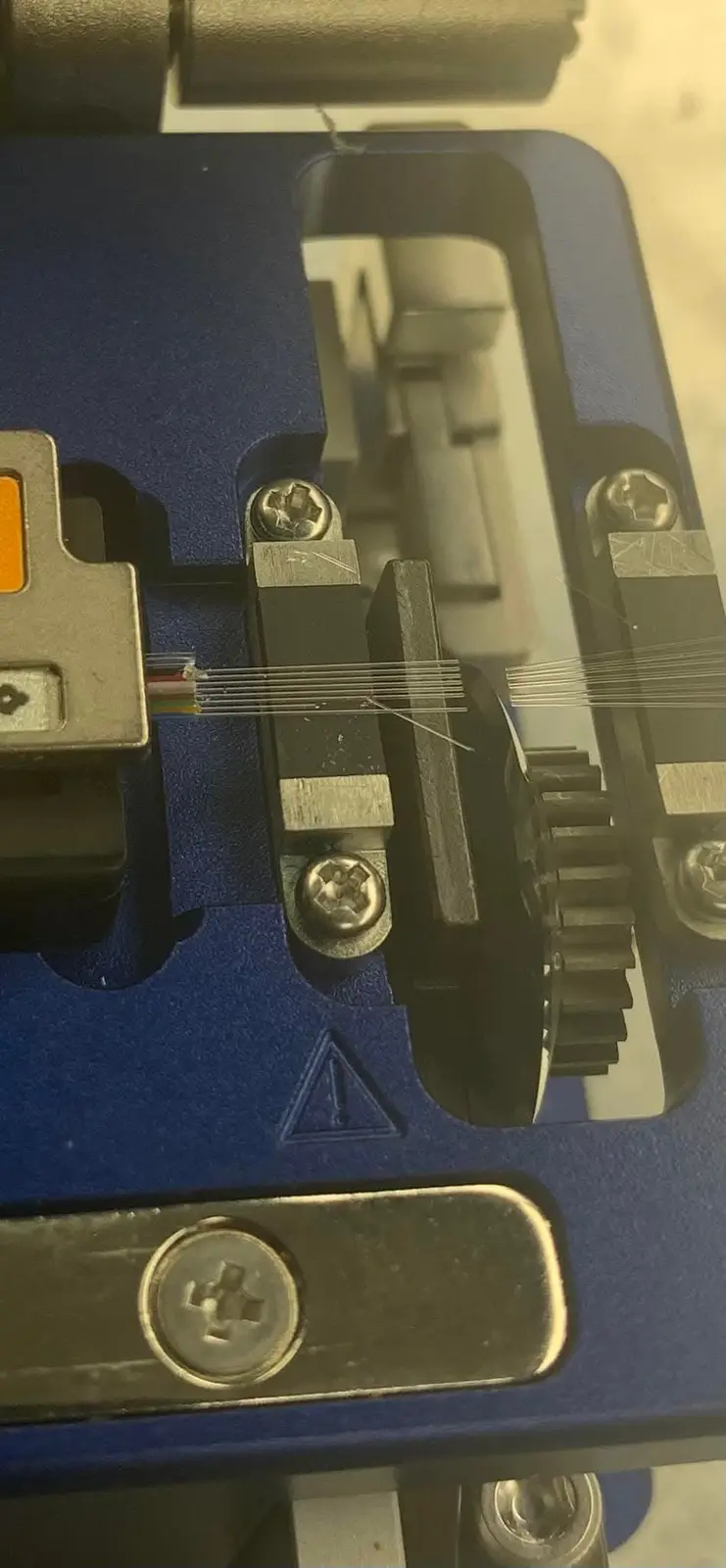 A close-up photo of a fiber optic cable being stripped by a blue industrial tool, showing the thin glass fibers and metal…