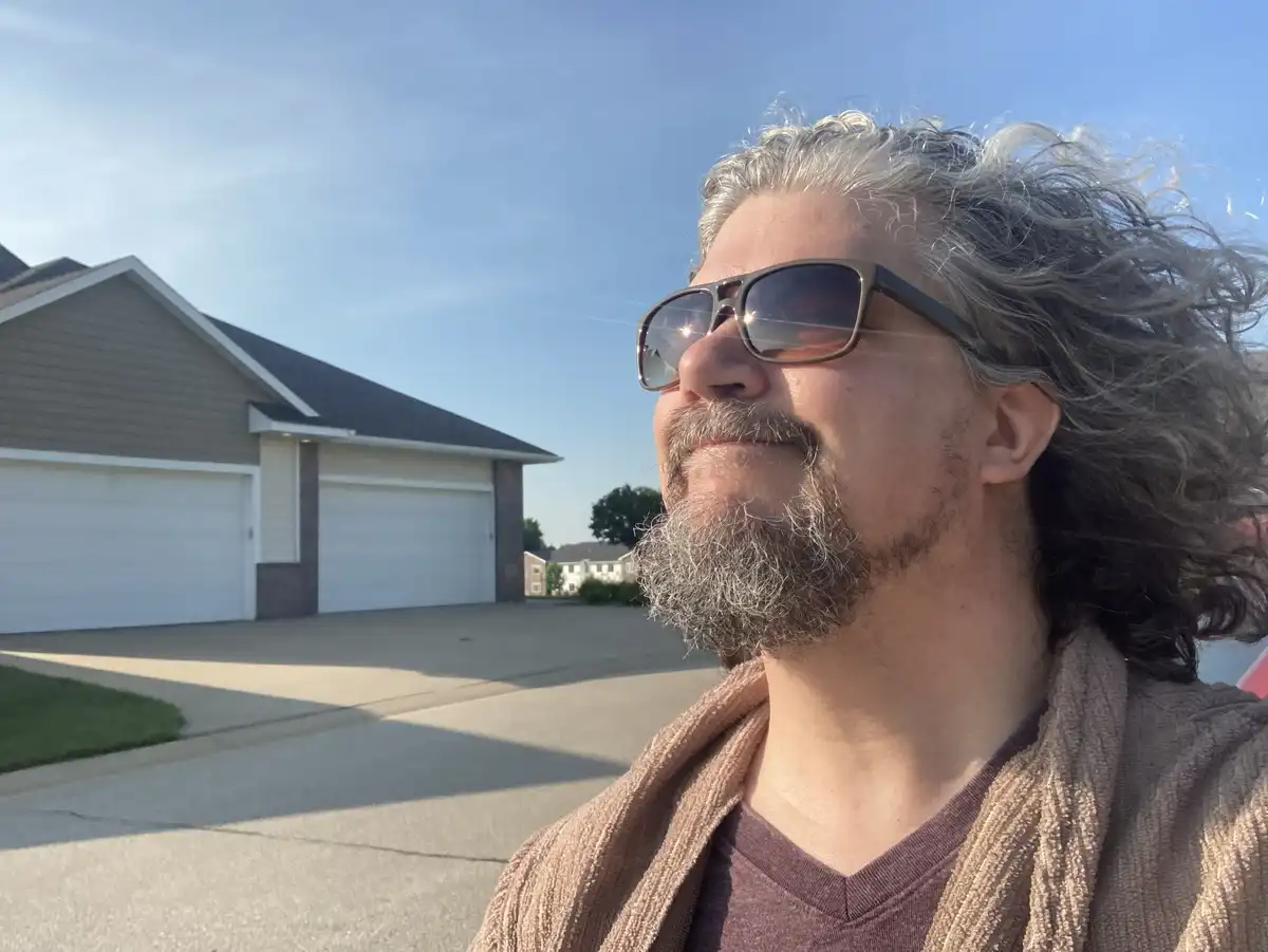 A man with long gray hair and a beard, wearing sunglasses and a brown robe, stands outside a suburban home with a satisfied…
