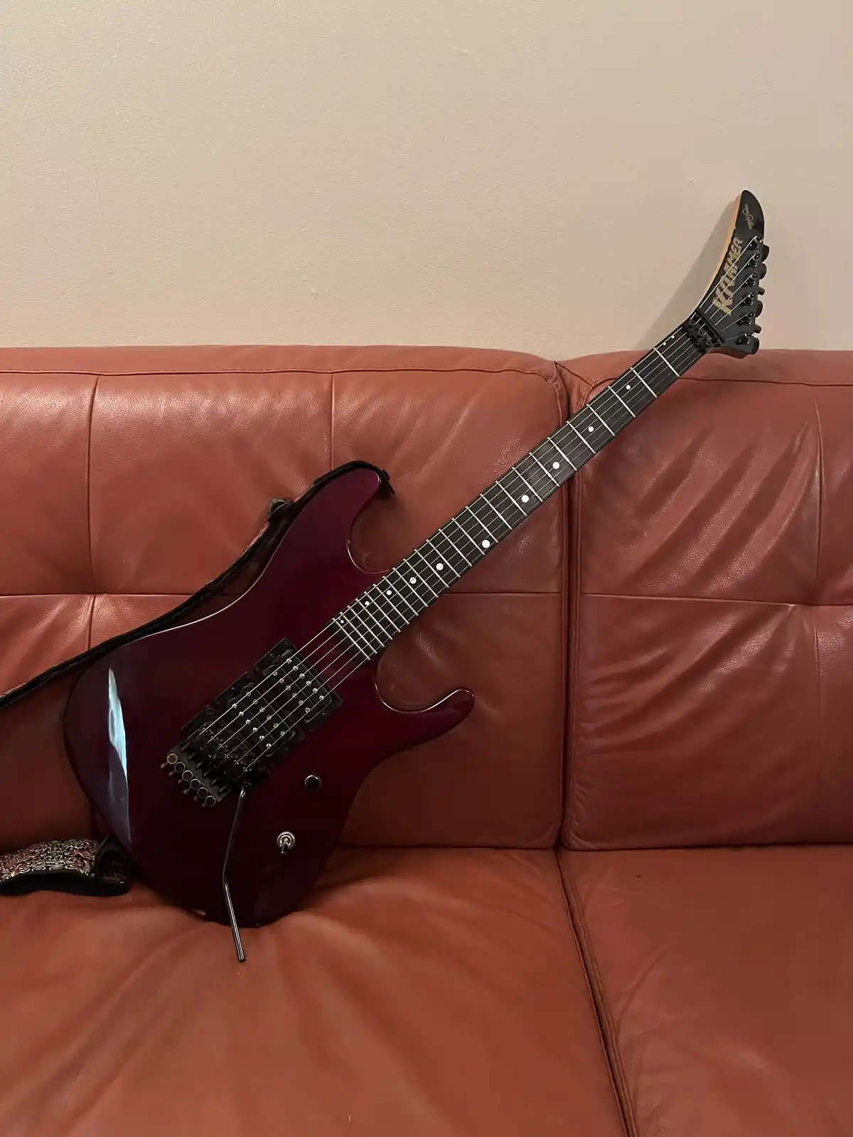 A glossy, deep red electric guitar with a black fretboard and headstock rests diagonally across a brown leather couch, its…
