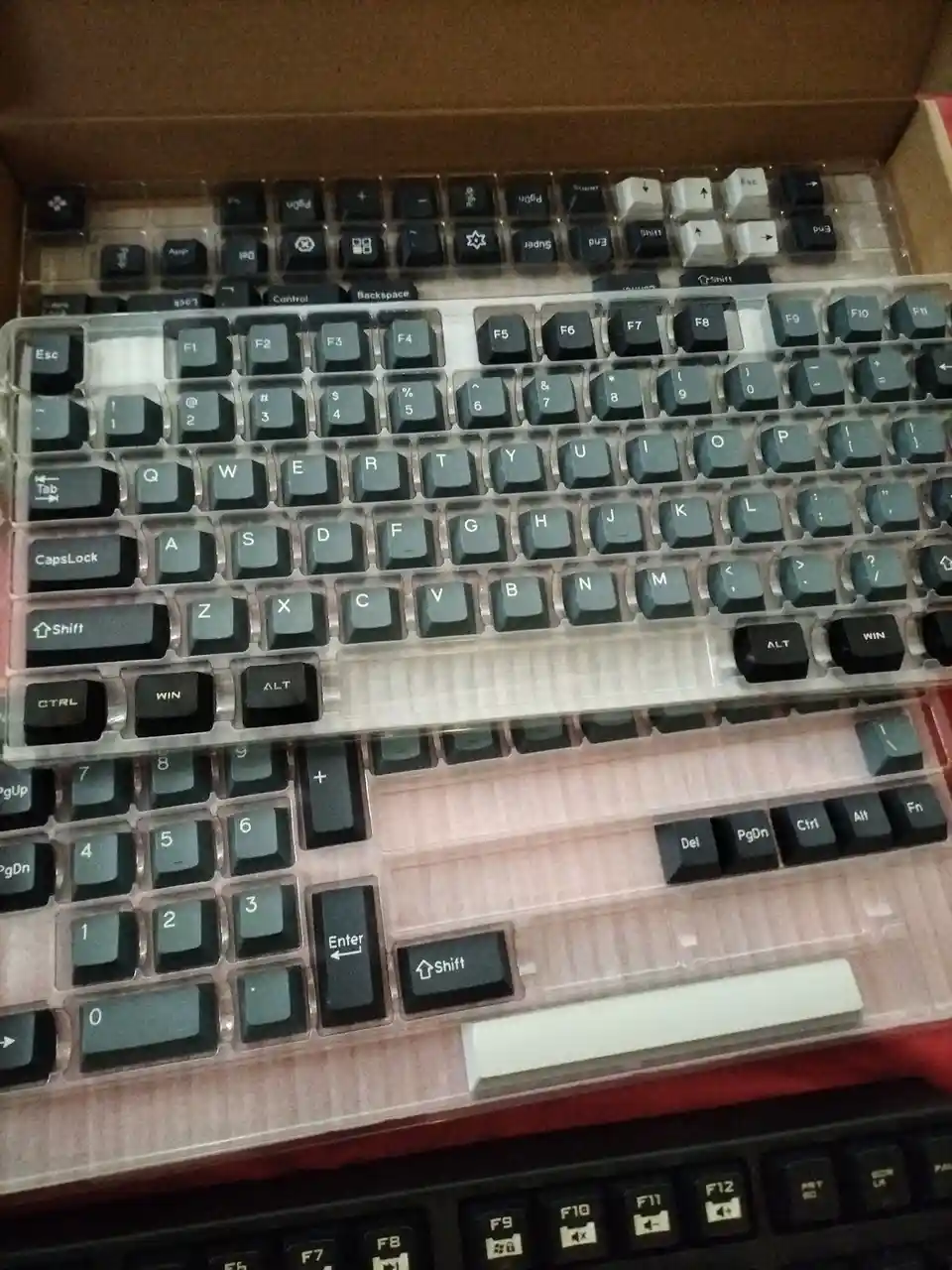 A close-up photo shows a mechanical keyboard with its keycaps still in the transparent plastic tray, sitting inside its…