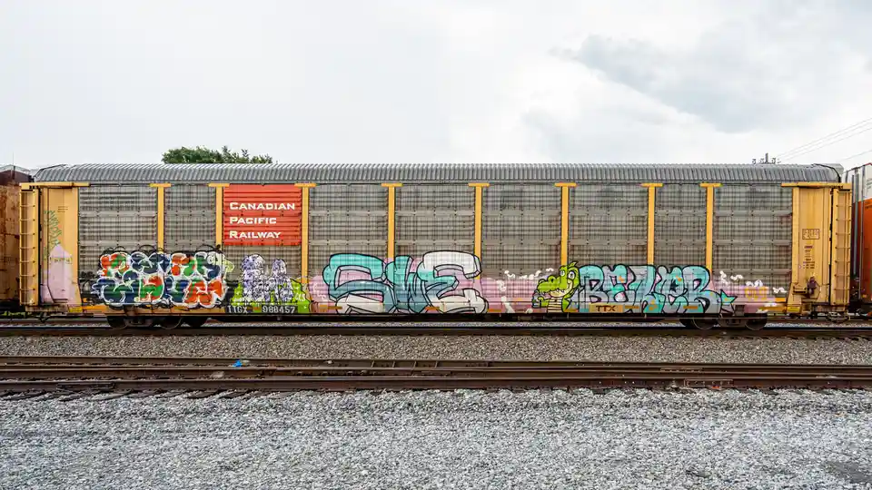 A side view of a Canadian Pacific Railway freight car covered in colorful graffiti tags, parked on train tracks under a…