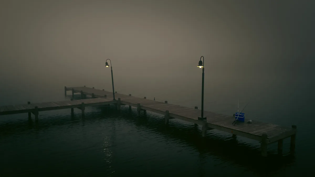 A foggy, dimly lit wooden dock extends over dark water, with two illuminated lamps and a blue cooler with fishing rods on…