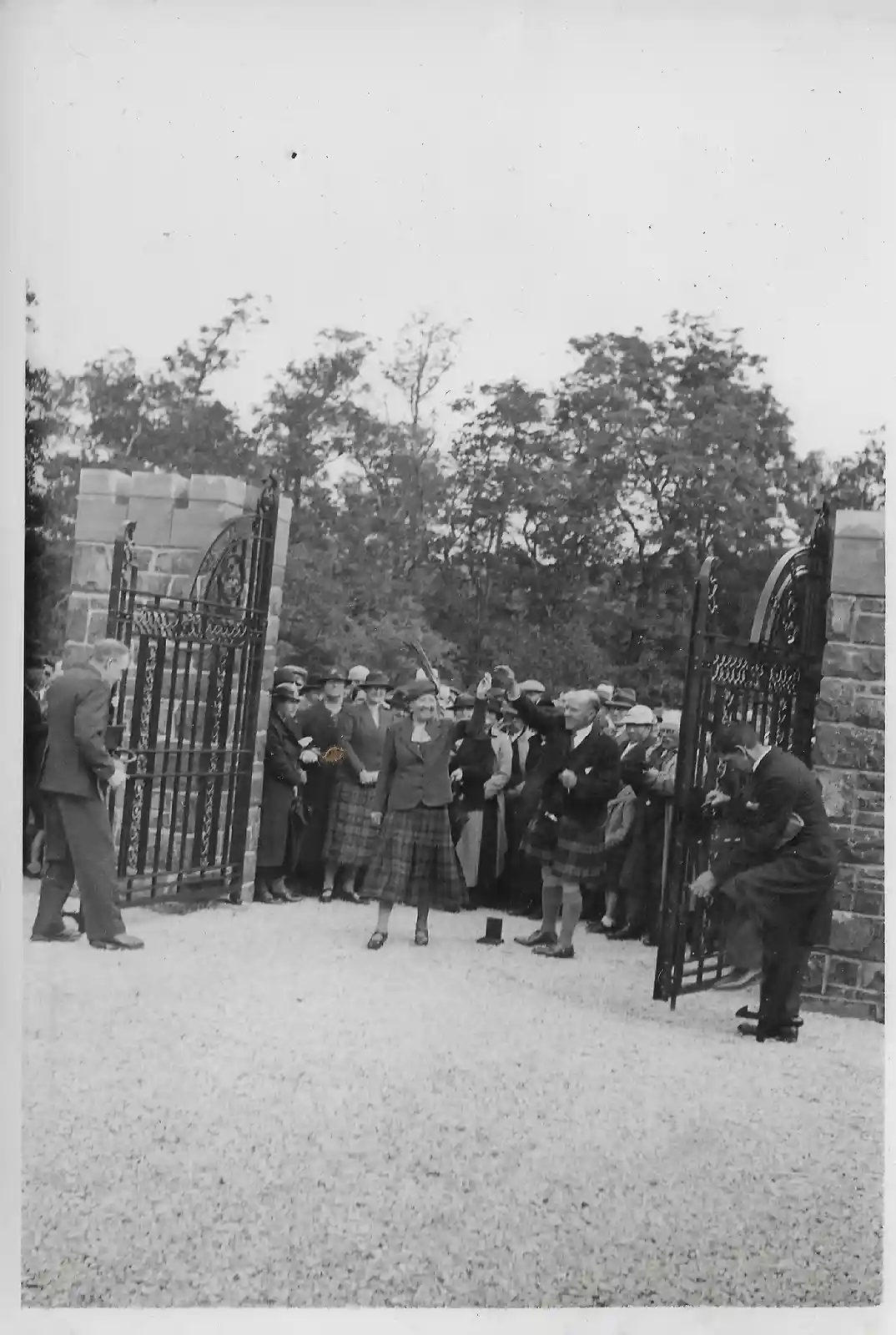 A black-and-white historical photo shows a woman in a plaid skirt and hat standing in front of ornate gates, smiling and…