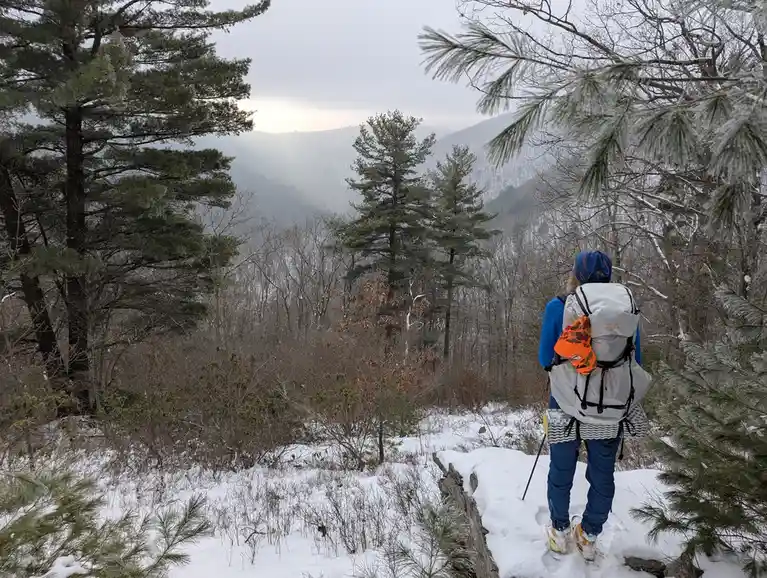 A person wearing a large backpack, blue jacket and pants, and a blue hat stands on a snowy trail overlooking a forested…