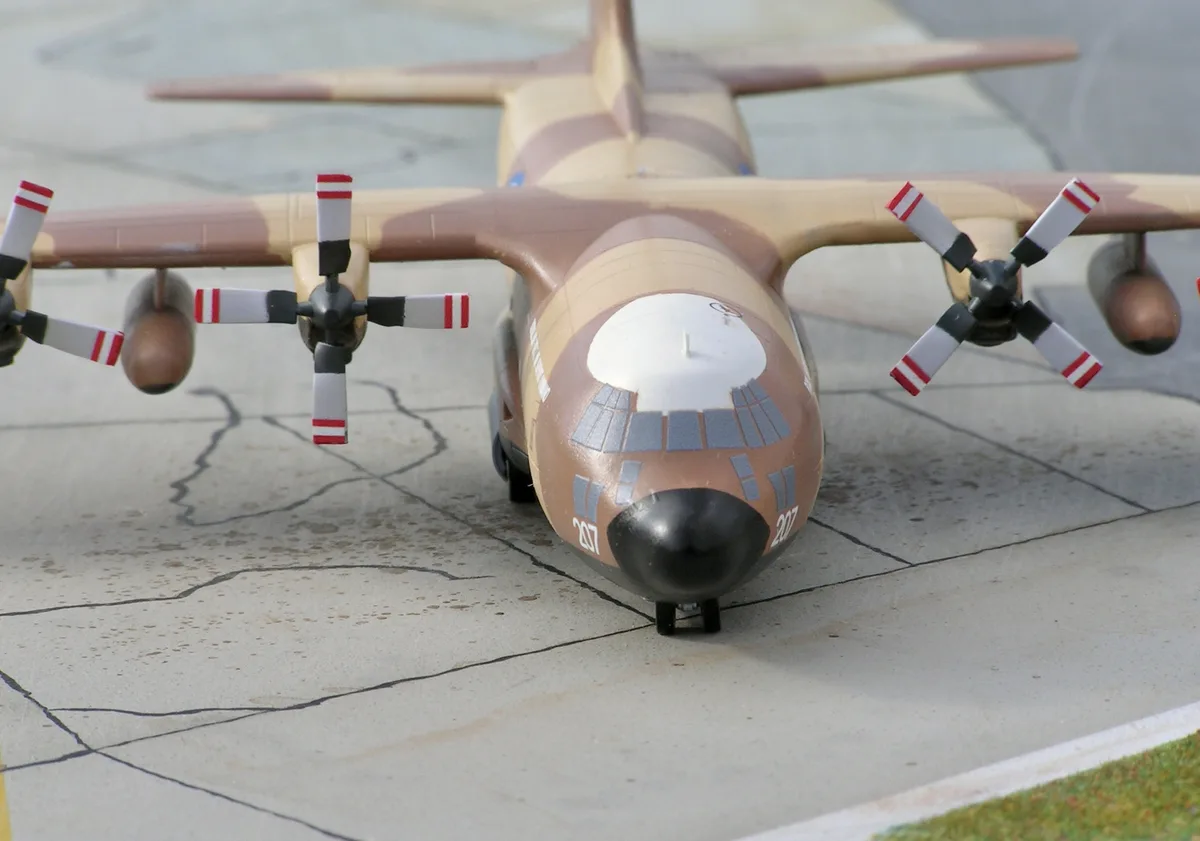 A close-up photo of a detailed model airplane with a desert camouflage paint scheme, four propellers with red and white…