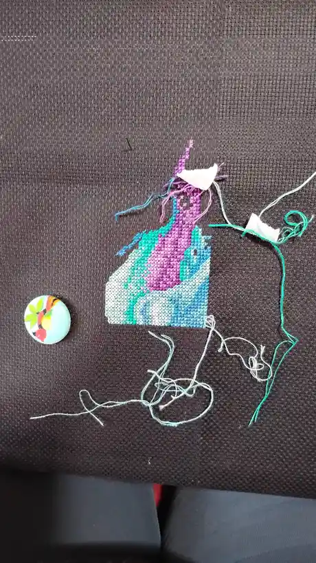 A partially completed cross-stitch project on dark fabric shows a colorful unicorn figure with loose threads and tangled…