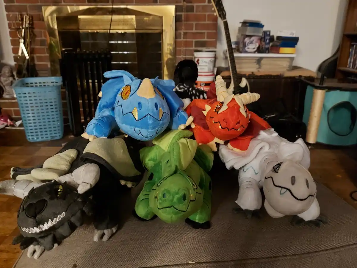 A collection of five plush dragons in various colors—blue, red, green, black, and white—are arranged together on a couch in…