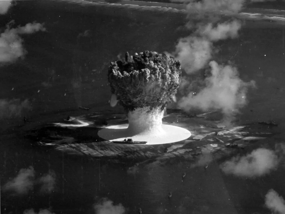 A black-and-white historical photograph shows a massive nuclear mushroom cloud rising over water, with ships visible in the…