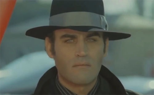a close-up of a man wearing a black fedora with a silver band and a dark coat, looking intensely at the camera with a…