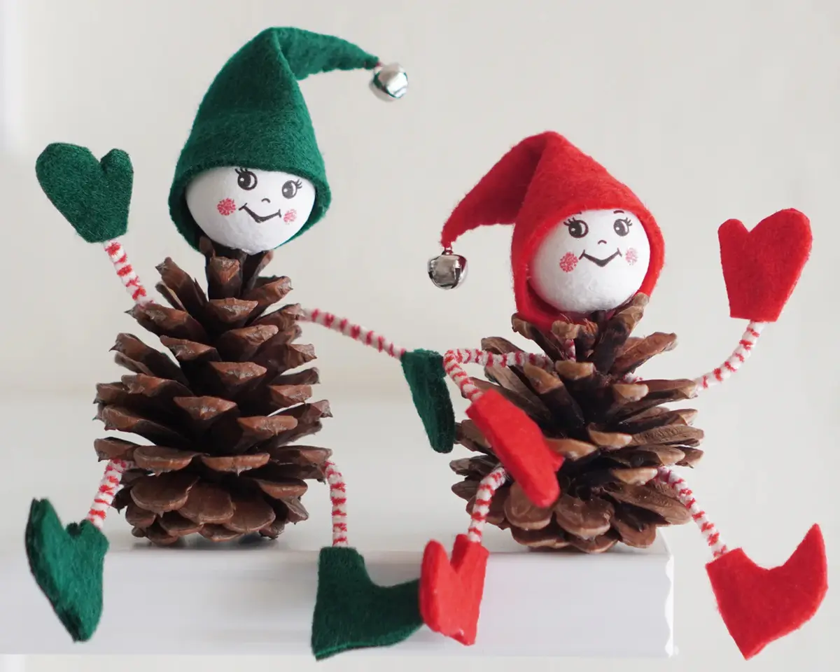Two handmade pinecone elves, one in a green hat and one in a red hat, are holding hands with heart-shaped mittens. Each has…