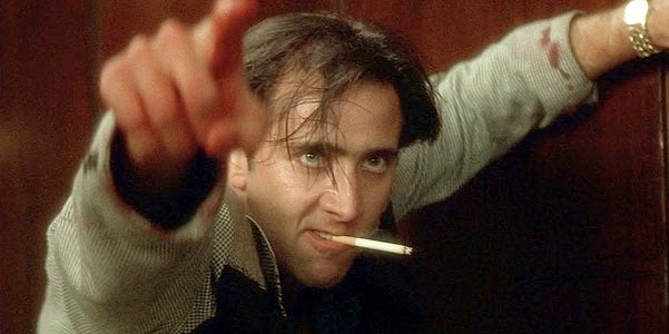 a man with messy hair and a cigarette in his mouth points directly at the viewer while leaning against a wooden surface; his…