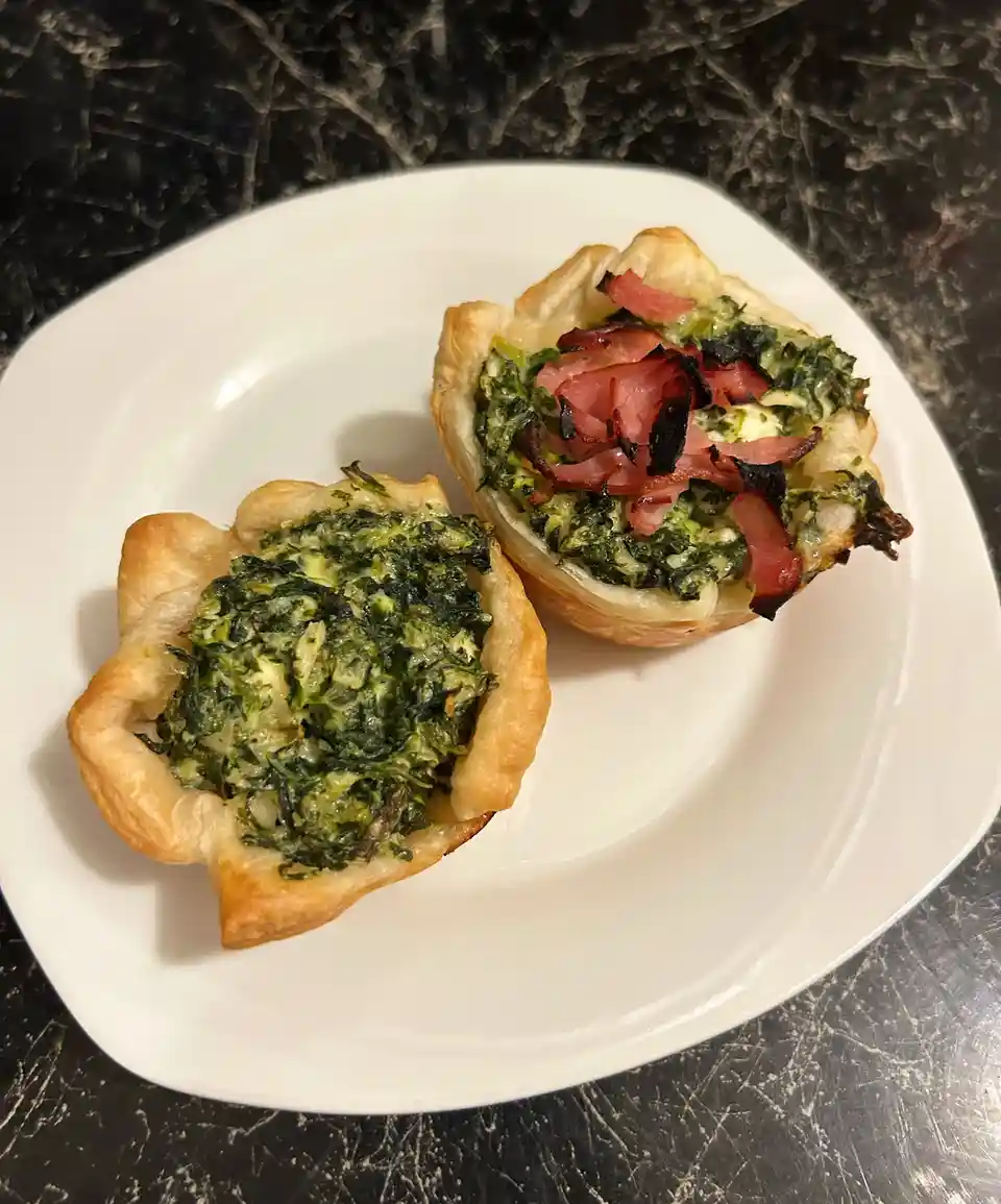 Two small, golden-brown pastry cups sit on a white plate, one filled with spinach and cheese filling, the other identical…