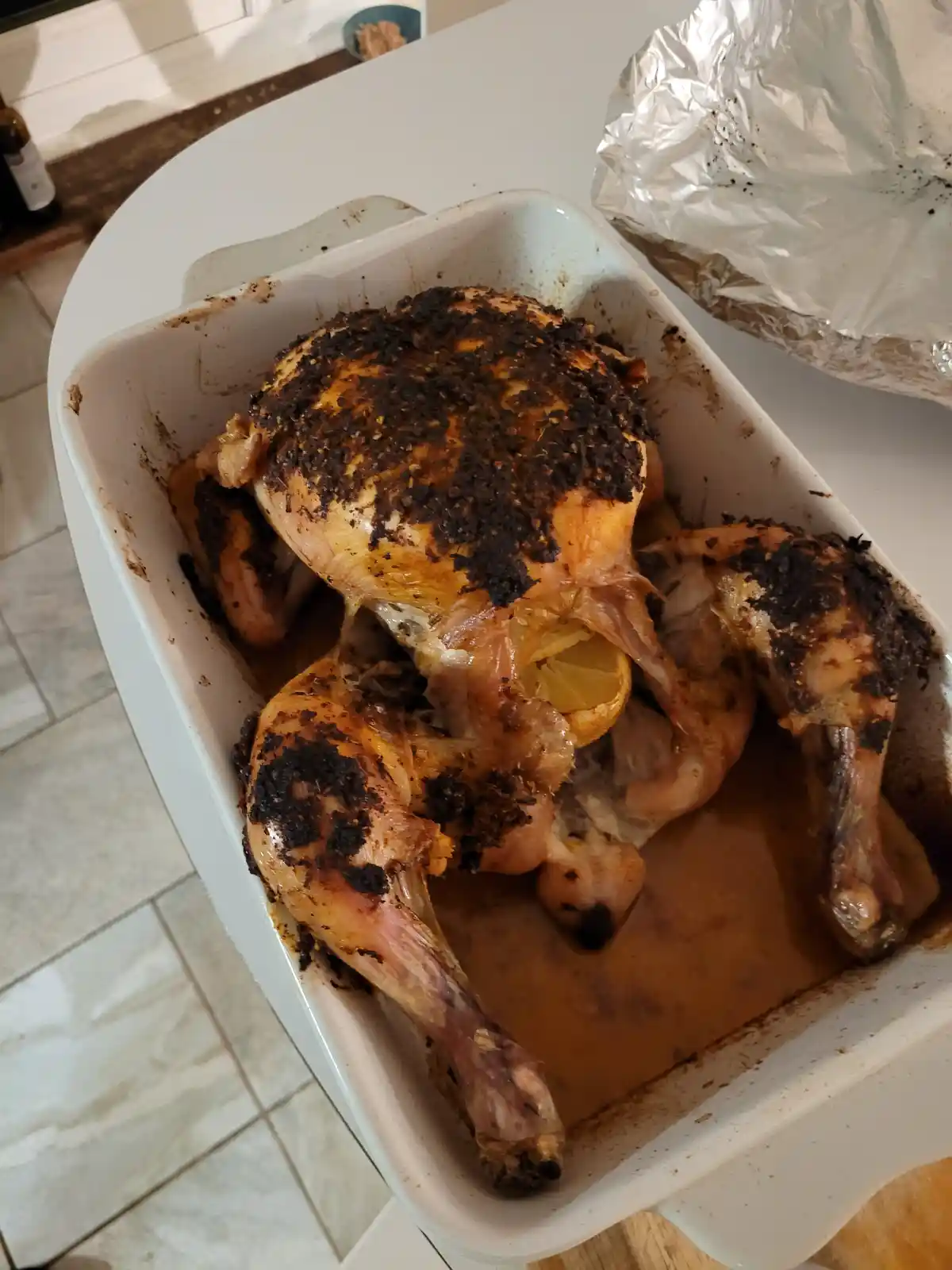 A roasted chicken in a white baking dish has severely burnt, charred seasoning on its skin, with a lemon wedge visible…