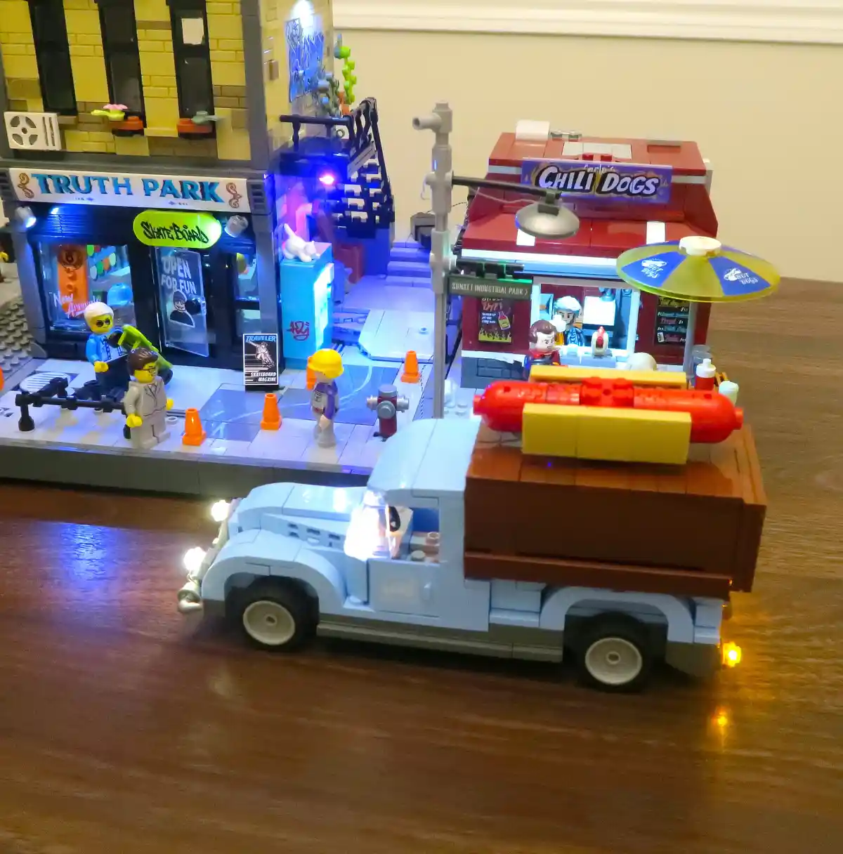 A detailed LEGO diorama depicts a street scene with a blue vintage truck carrying a giant red hot dog on a yellow bun in its…