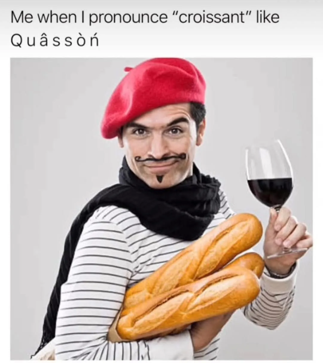 A man wearing a red beret, black scarf, and striped shirt holds two baguettes in one arm and a glass of red wine in the…