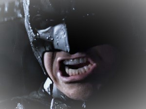 A close-up of Batman's face, partially obscured by his cowl, with raindrops visible on the mask and his mouth open in a…