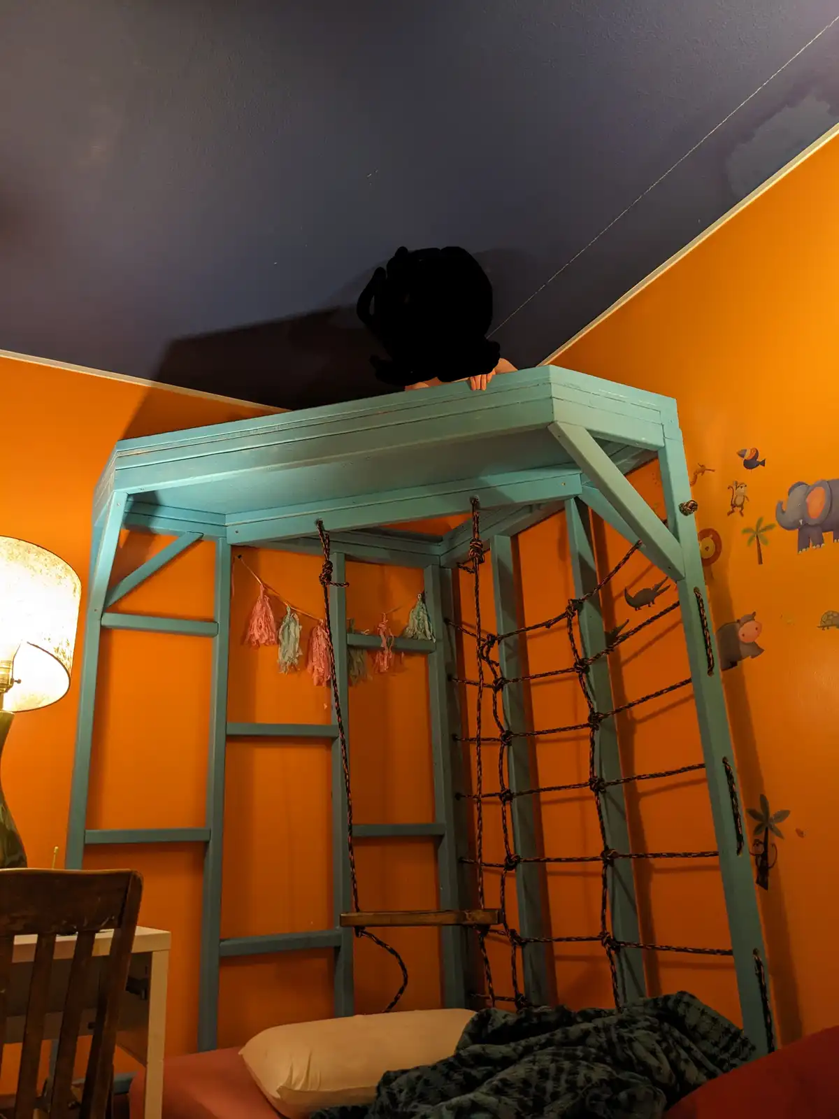 A child with their face obscured by a black blur is perched on the top edge of a teal jungle gym-style bunk bed in a…