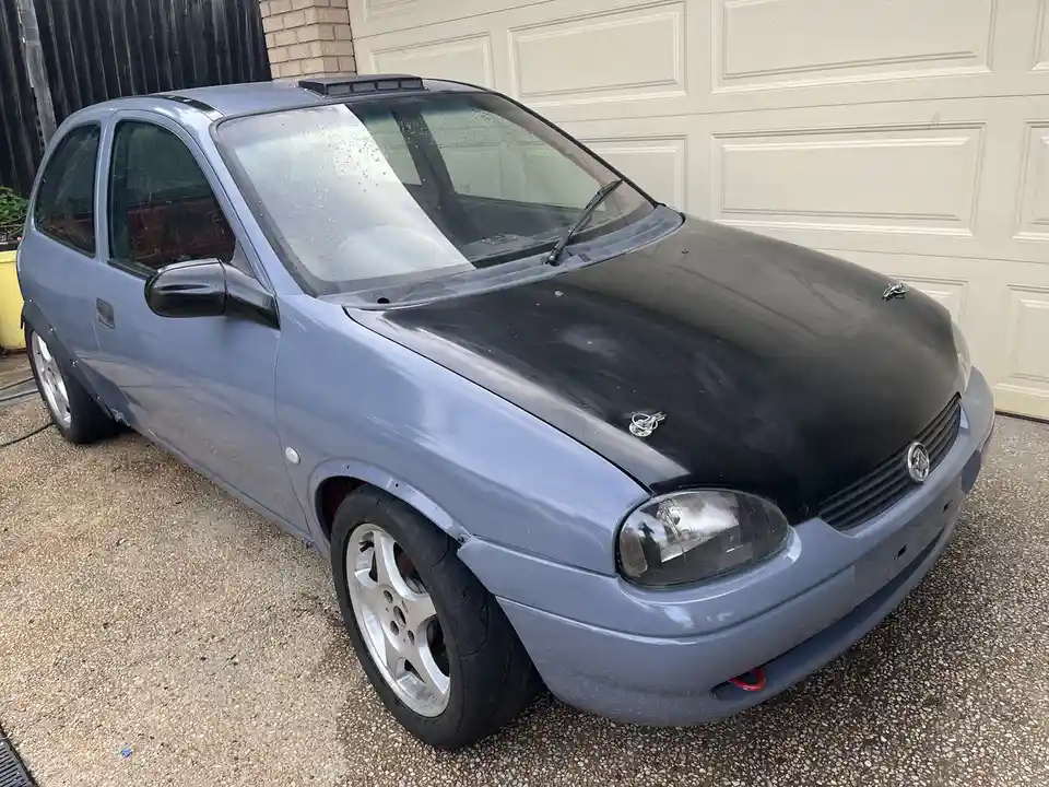 A light blue Holden Barina hatchback is parked in front of a white garage door, showing visible modifications including a…