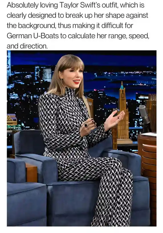 Taylor Swift is seated in a blue armchair on a talk show set with a nighttime cityscape backdrop. She wears a…