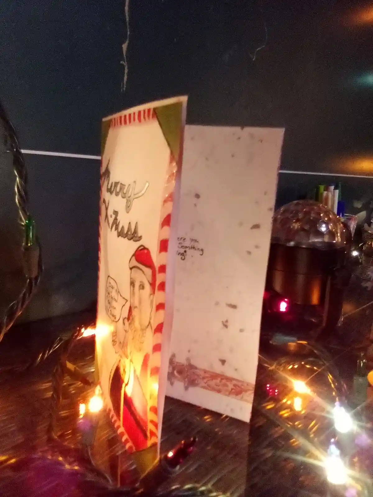 A handmade Christmas card stands upright on a table surrounded by string lights, its front decorated with a Santa Claus…