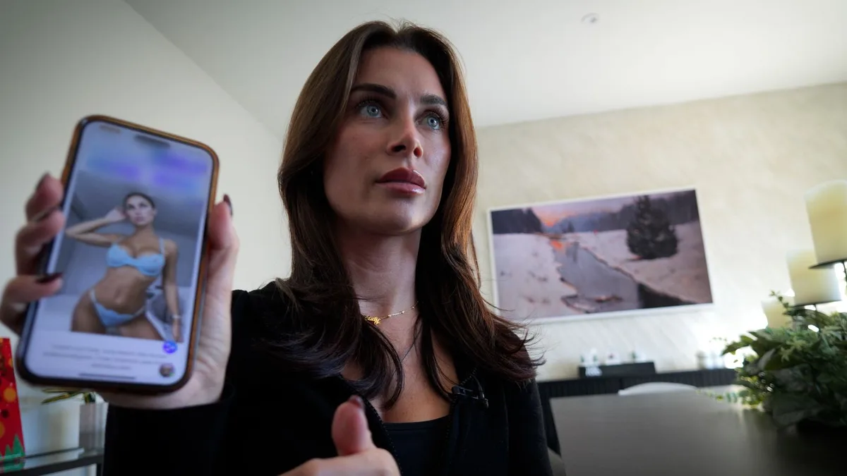 A woman with brown hair and blue eyes looks upward with a shocked expression while holding up a smartphone displaying a…