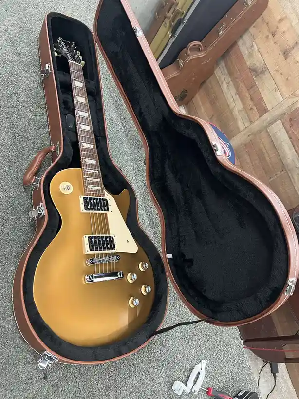 A golden electric guitar sits inside an open brown hardshell case on a gray carpet, with tools and a screwdriver visible…