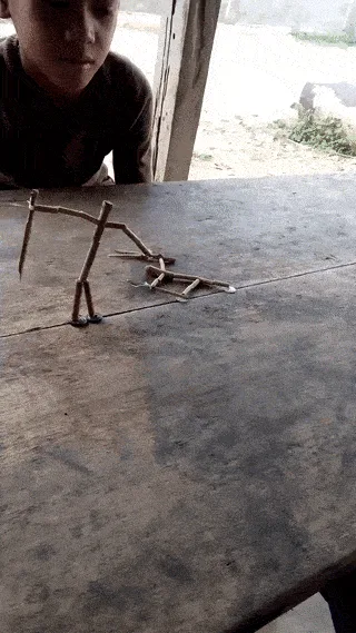 A young boy sits at a wooden table, intently watching a small stick figure he built collapse and reassemble in a sequence of…