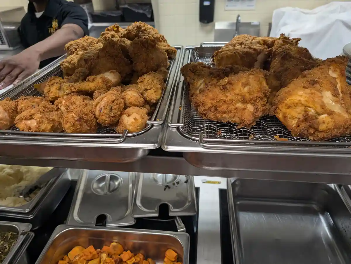 Two metal trays filled with golden-brown fried chicken pieces sit on a buffet line, with a worker's arm visible in the…