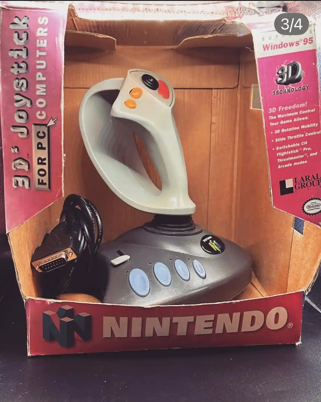 A gray and black 3D joystick controller sits inside a cardboard box that prominently displays the Nintendo logo on the…
