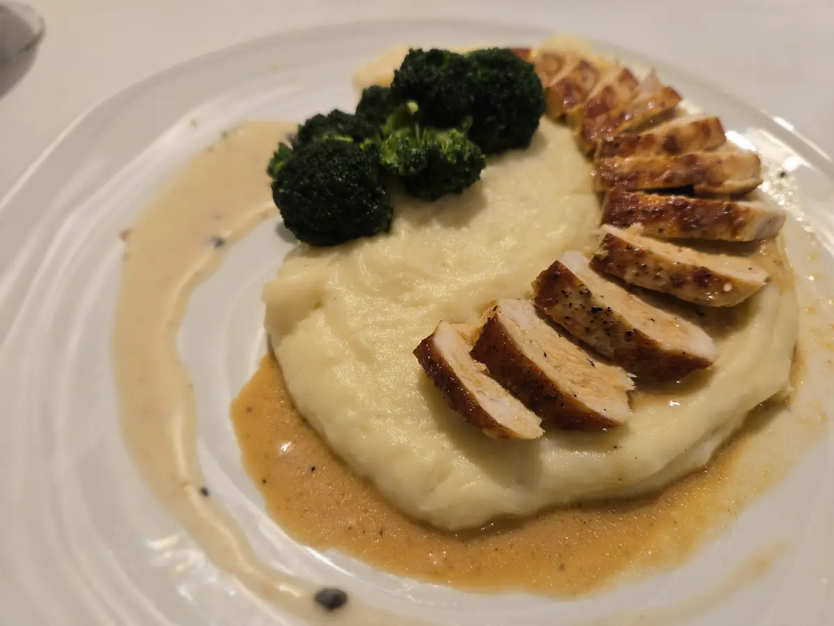 A plated meal featuring sliced grilled chicken breast atop a mound of creamy mashed potatoes, accompanied by steamed…