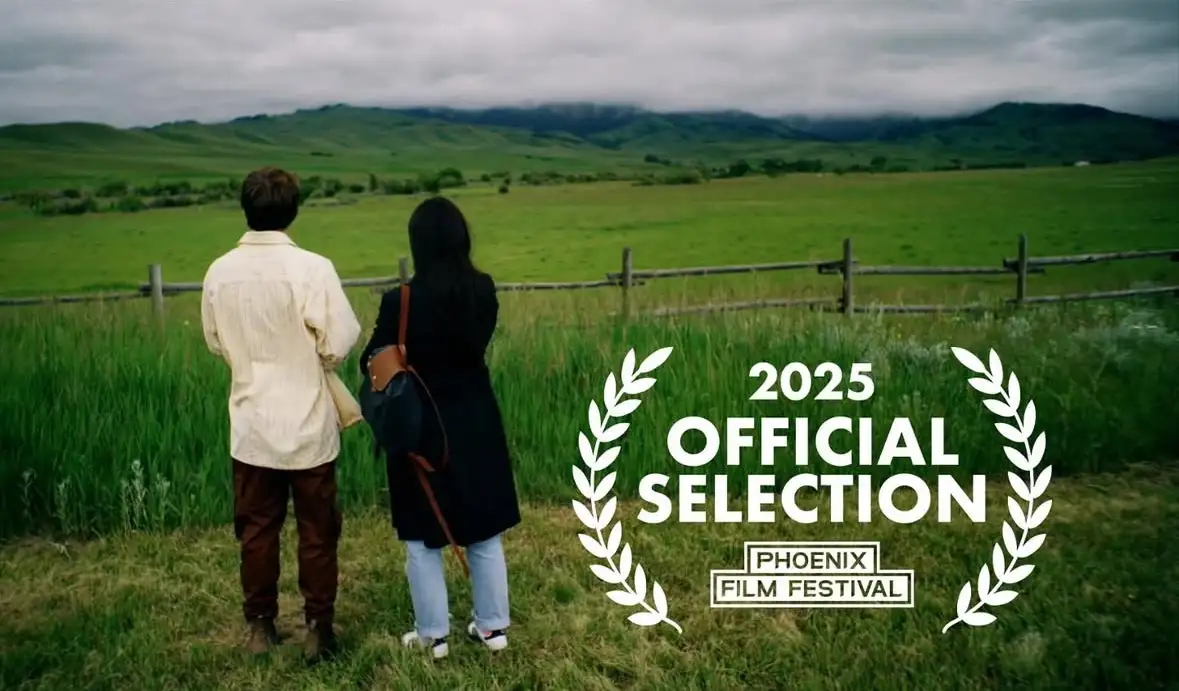 Two people stand back-to-back in a grassy field, looking out at rolling green hills under a cloudy sky. A white laurel…