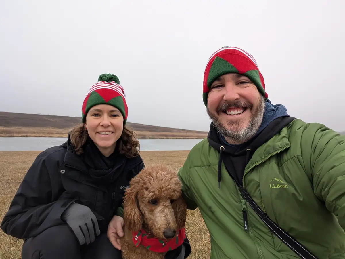 A man and woman in festive red-and-green elf hats pose for a selfie outdoors with a brown poodle wearing a matching holiday…