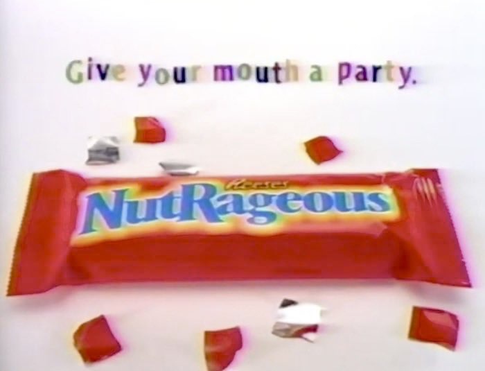 A red-wrapped Reese's NutRageous candy bar is centered on a white background with scattered red and silver foil pieces…