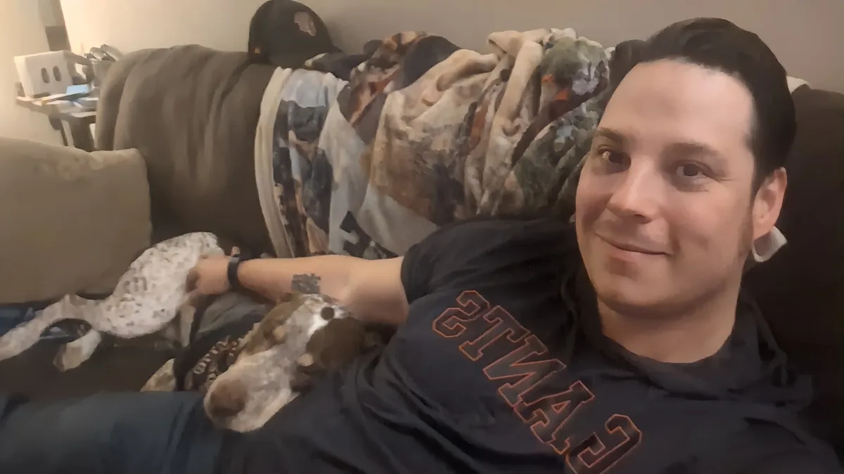 a man in a dark hoodie with 'STARS' written in orange letters lies on a couch, smiling at the camera while holding a spotted…