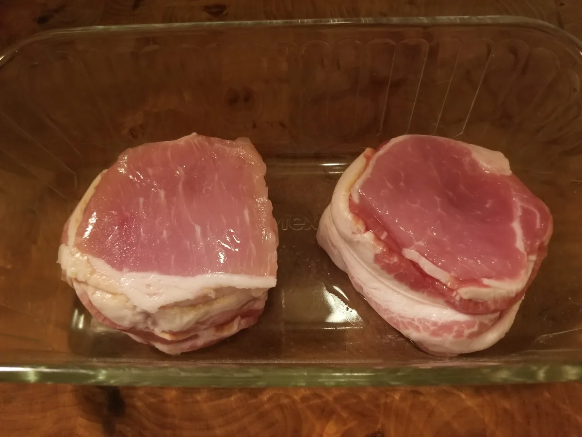 Two stacks of raw bacon sit side-by-side in a clear glass baking dish on a wooden surface. The bacon on the left is a…